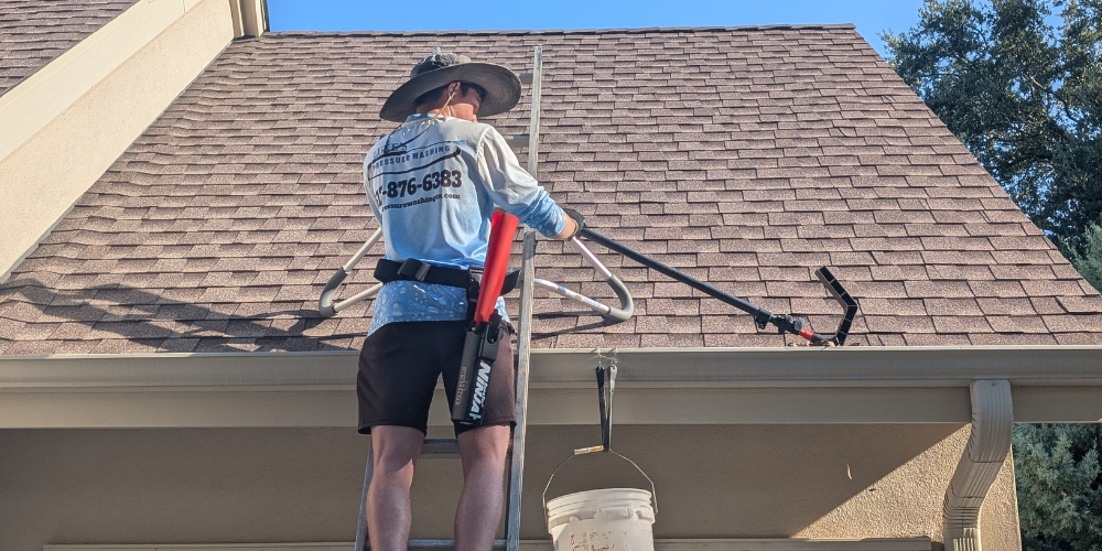 Luke’s Pressure Washing technician cleaning gutters safely on ladder using hand tools and bucket system for residential roof maintenance in Georgetown, Texas.